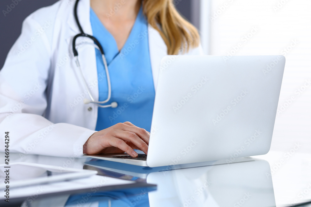 Doctor typing on laptop computer while sitting at the glass desk in hospital office. Physician at work. Medicine and healthcare concept Doctor typing on laptop computer while sitting at the glass desk in hospital office. Physician at work. Medicine and healthcare concept