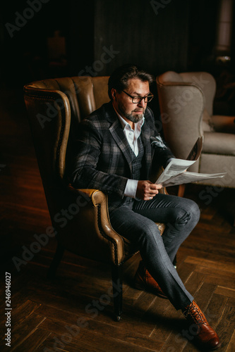 Portrait of mature successful caucasian fashion designer wearing elegant suit and glasses, reading article about his new apparel collection in newspaper with thoughtful concentrated look