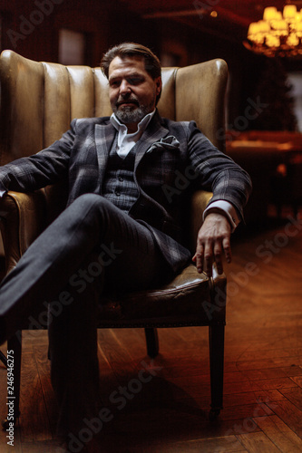 Portrait of wealthy prosperious handsome businessman, dressed in tailored three-piece suit sitting in leather arm-chai at elite gentlemen club with lighted multi-lamp chandelier behind.