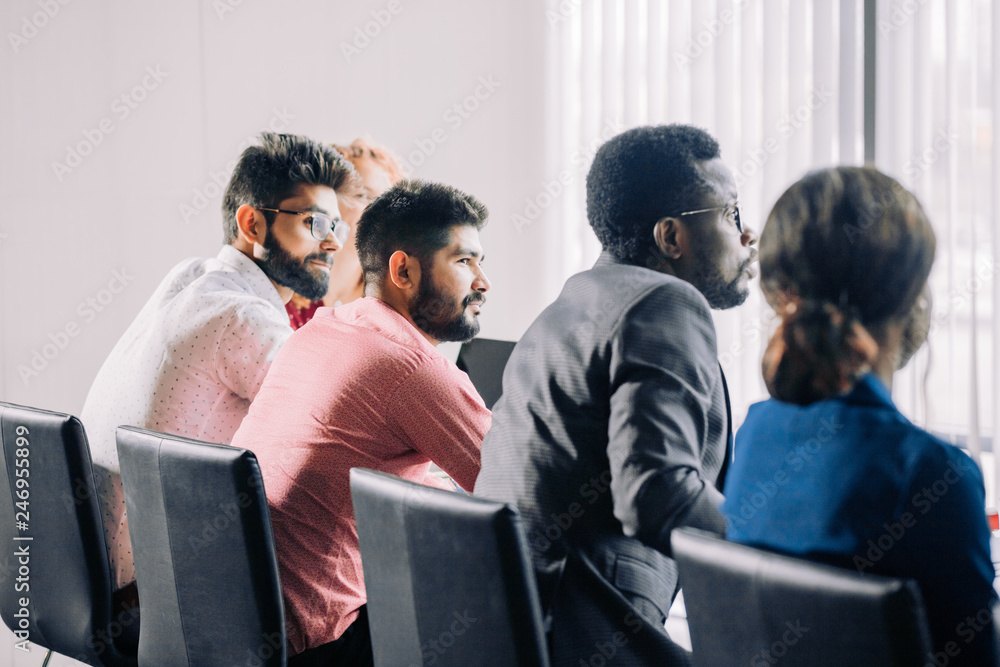 Backside of group of diverse multiracial applicants for a vacant post or corporate job sitting in a long line before HR specialist explaining test task over panoramic windows background. Backside of group of diverse multiracial applicants for a vacant post or corporate job sitting in a long line before HR specialist explaining test task over panoramic windows background.