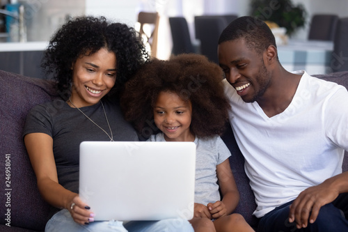 Happy african american family with child girl having fun using laptop together at home, black parents and little kid daughter watch funny video, do online shopping, make call look at computer screen