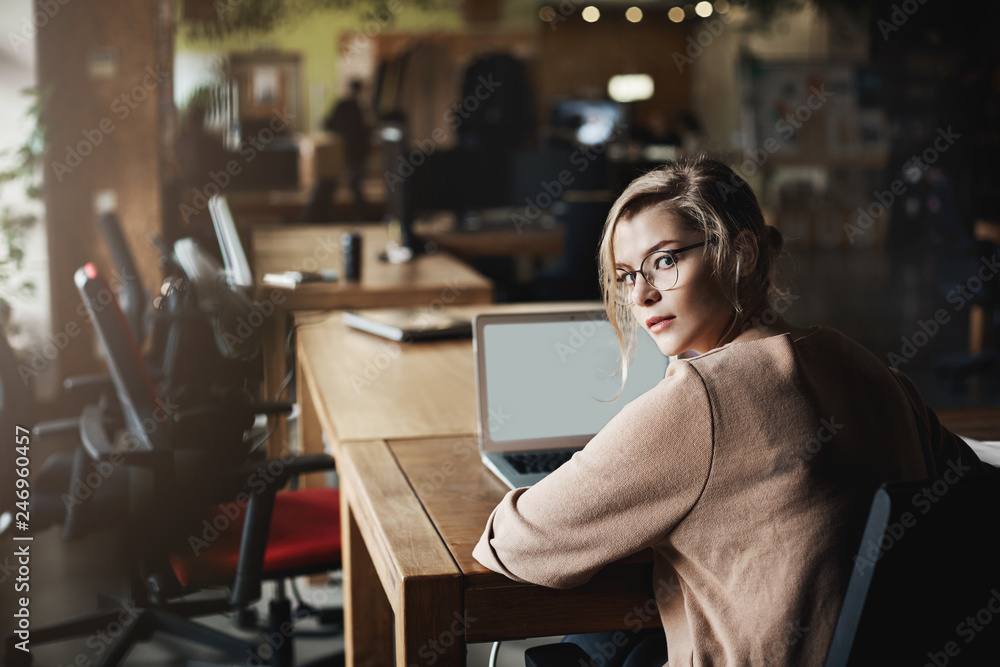 Attractive determined caucasian blond woman in glasses, turning back to look at camera, being called by coworker while sitting in office, working via laptop, communicating with customers