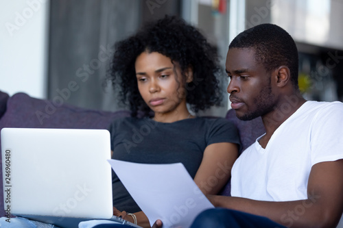 Serious african american couple reading document letter pay domestic bills checking bank account online on laptop at home  black family holding paper planning budget money expenses with computer app