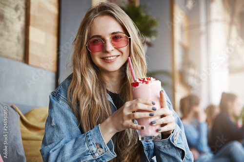 Take pause and drink tasty cocktail. Outdoor shot of carefree cute blond female student in stylish pink sunglasses, holding drink with strawberry and gazing with caring smile at camera