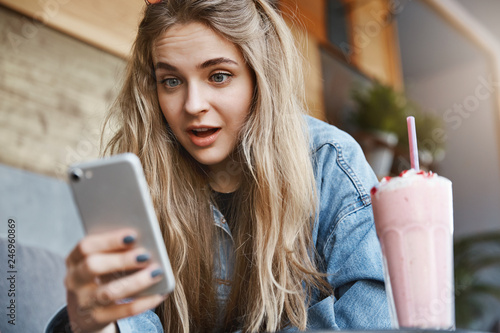 Portrait of stunned and impressed attractive blond woman in denim jacket, sitting in cafe and drinking strawberry cocktail, holding smartphone and dropping jaw from show, staring at gadget screen