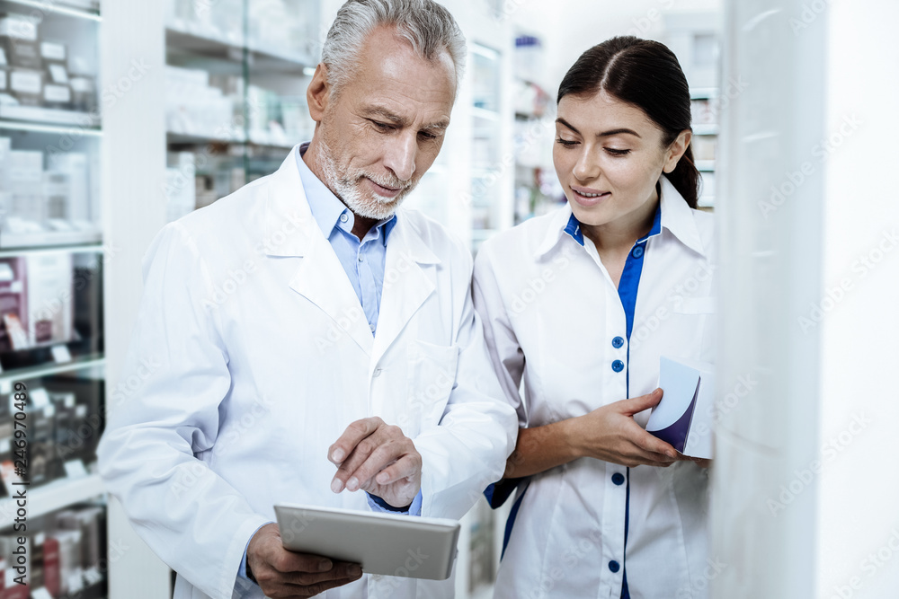 Mature bearded man in a white coat discussing medicines with his young colleague Mature bearded man in a white coat discussing medicines with his young colleague