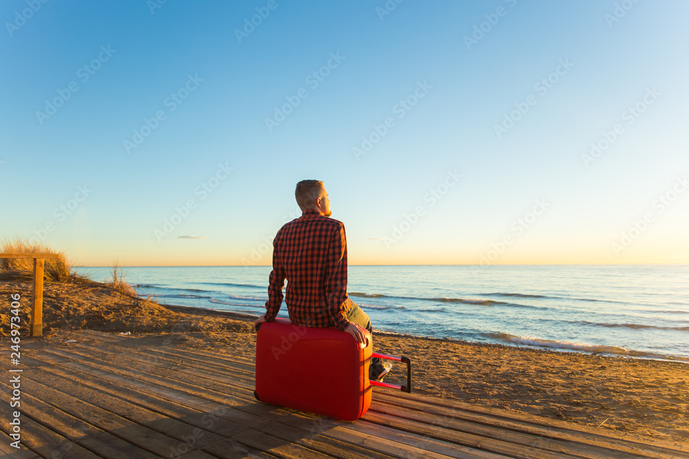 Fototapeta premium Holiday, travel and summer vacation concept -a man sitting on red suitcase and watching the sunset