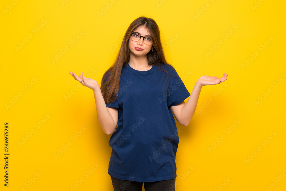Young woman with glasses over yellow wall having doubts while raising hands and shoulders Young woman with glasses over yellow wall having doubts while raising hands and shoulders