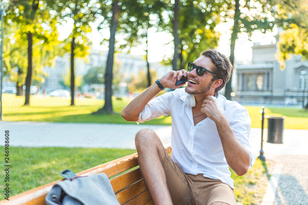 Cheerful man looking at side with toothy smile while talking to friend on smartphone and sitting on park bench.