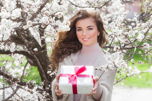 Young pretty girl holding present box outdoors. Woman on spring background with gift. Beautiful lady portrait. photo