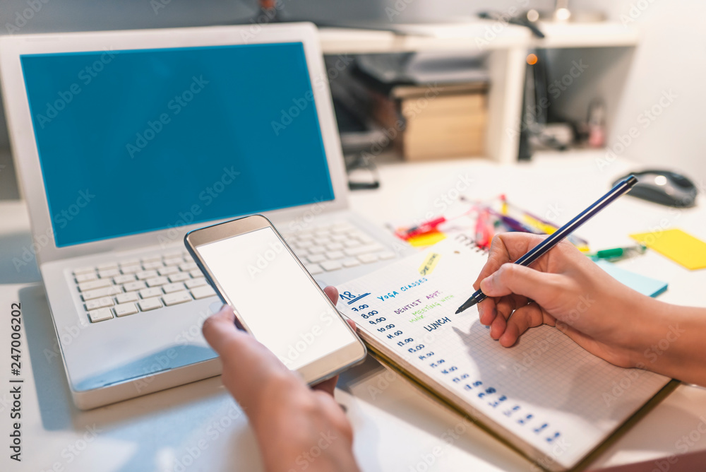 Woman hand writing in agenda consulting a mobile phone on a desk at home or office Woman hand writing in agenda consulting a mobile phone on a desk at home or office