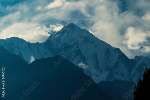 View on Himalayas from Annapurna Circuit Trek, Nepal. Clouds covered with clouds. Sharp slopes. Smaller mountain in front, covered in shadow. Overcast but sunny. High altitude climbing.