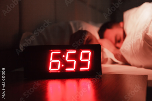 Rectangular alarm clock on the bedside table with red numbers, sleeping man in bed in dark room. Concept chef sleeping after hard day at restaurant, sleep disturbance, oversleep work, night shift