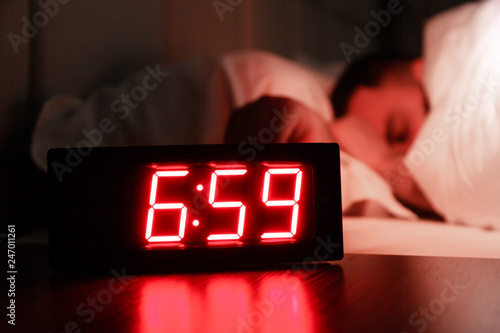 Rectangular alarm clock on the bedside table with red numbers, sleeping man in bed in dark room. Concept chef sleeping after hard day at restaurant, sleep disturbance, oversleep work, night shift