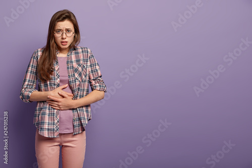 Painful feelings concept. Upset woman feels hungry, keeps hands on belly, suffers from pain, wears checkered casual shirt, transparent glasses, isolated on lilac studio wall. Bellyache concept photo