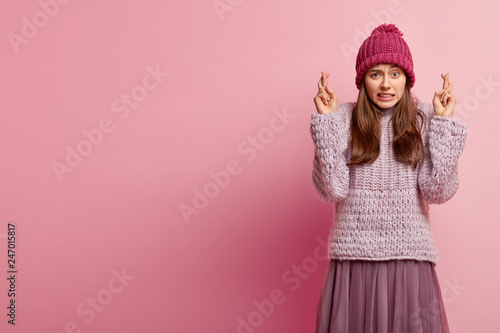 Photo Studio shot of hopeful woman has worried expression, keeps fingers crossed, believes in good fortune, stands over pink studio wall with empty space on left side for your promotional content