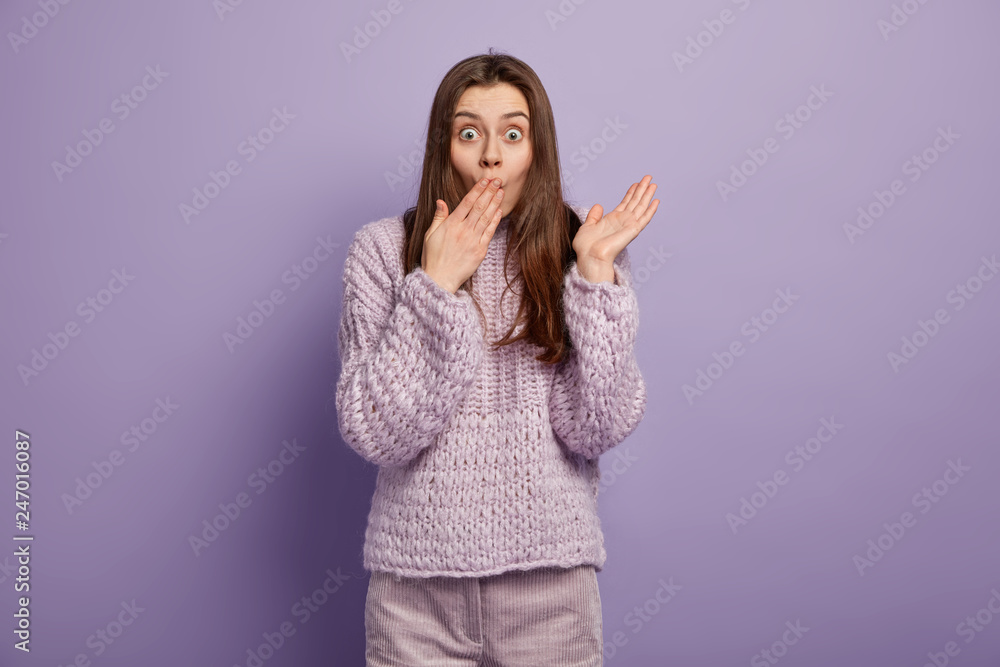 Potrait of attractive woman covers mouth with palm, hears shocking news, spreads palm, wears purple warm jumper in one tone with background, expresses disbelief. People and astonishment concept Potrait of attractive woman covers mouth with palm, hears shocking news, spreads palm, wears purple warm jumper in one tone with background, expresses disbelief. People and astonishment concept