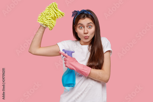 Indoor shot of surprised European housewife points at muscle, carries sponge and cleanser, wears headband, brings house to order, isolated over pink background. Cleaning and housework concept photo