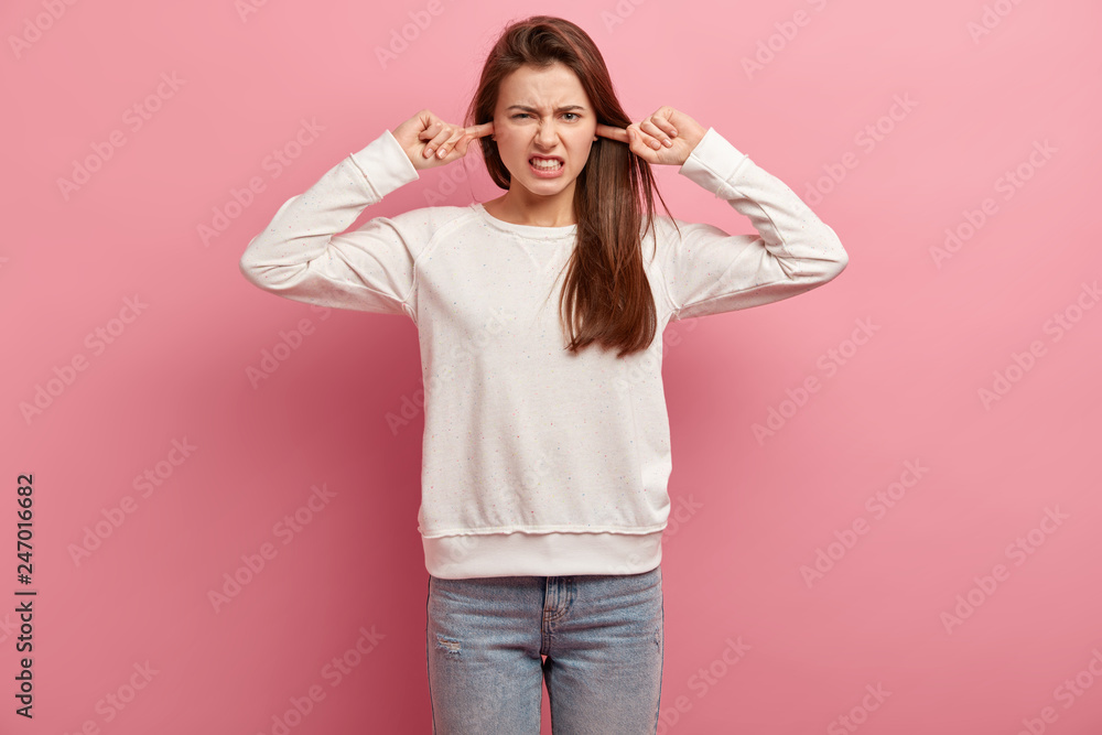Image of displeased young lady plugs ears, clenches teeth, hears noisy irritated sound, dressed in casual white jumper and jeans, stands against pink background, ignores hearing something annoying
