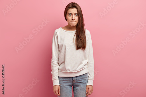 Puzzled young European woman presses lips, has bugged eyes, feels frustrated, wears casual sweatshirt and jeans, stands against rosy background, tries to make choice, dissatisfied with something