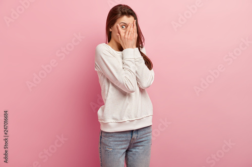 Fotografi Sideways shot of surprised woman covers face with palms, peeks through fingers,