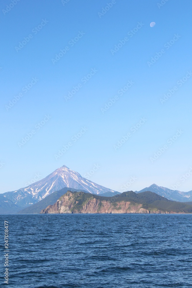 View of Vilyuchinsky volcano (also called Vilyuchik) from water. It's a stratovolcano in the southern part of Kamchatka Peninsula, Russia. Moon is visible in the sky.