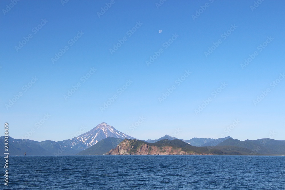 View of Vilyuchinsky volcano (also called Vilyuchik) from water. It's a stratovolcano in the southern part of Kamchatka Peninsula, Russia. Moon is visible in the sky. View of Vilyuchinsky volcano (also called Vilyuchik) from water. It's a stratovolcano in the southern part of Kamchatka Peninsula, Russia. Moon is visible in the sky.