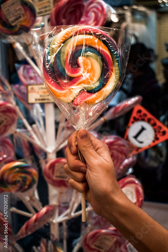 Female hand holding a rainbow colour colored lollipop in front of a holder with lollipops