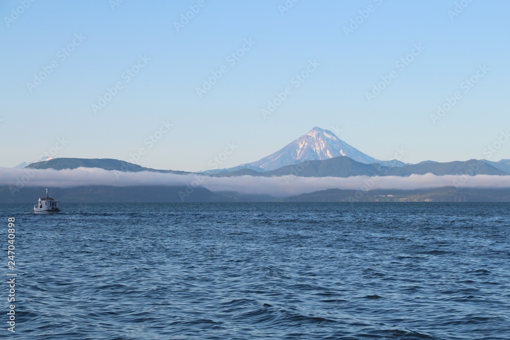 View of the Vilyuchinsky volcano (also called Vilyuchik) from tourist boat. The cloud lies on the coastal cliffs. Tourist boat is visible in the waters of Avacha Bay on the Kamchatka Peninsula, Russia View of the Vilyuchinsky volcano (also called Vilyuchik) from tourist boat. The cloud lies on the coastal cliffs. Tourist boat is visible in the waters of Avacha Bay on the Kamchatka Peninsula, Russia