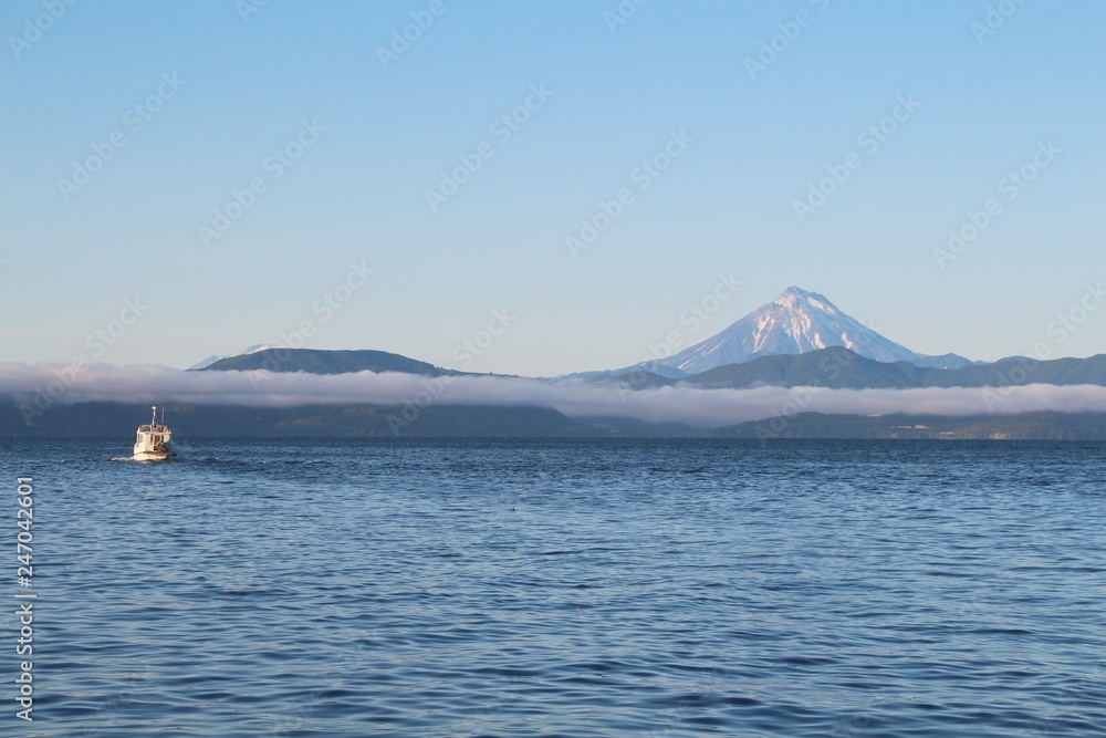 View of the Vilyuchinsky volcano (also called Vilyuchik) from tourist boat. The cloud lies on the coastal cliffs. Tourist boat is visible in the waters of Avacha Bay on the Kamchatka Peninsula, Russia