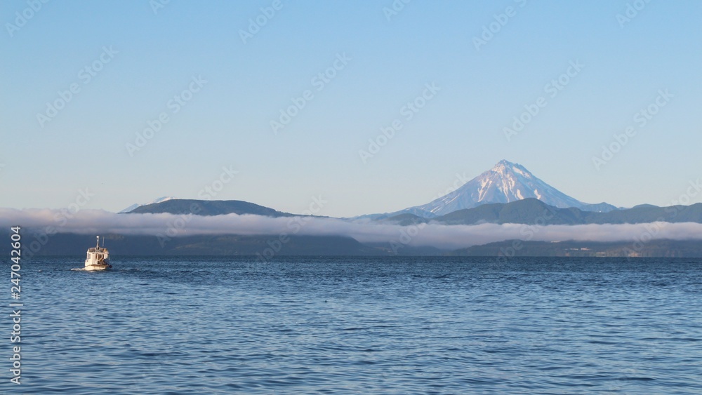 View of the Vilyuchinsky volcano (also called Vilyuchik) from tourist boat. The cloud lies on the coastal cliffs. Tourist boat is visible in the waters of Avacha Bay on the Kamchatka Peninsula, Russia