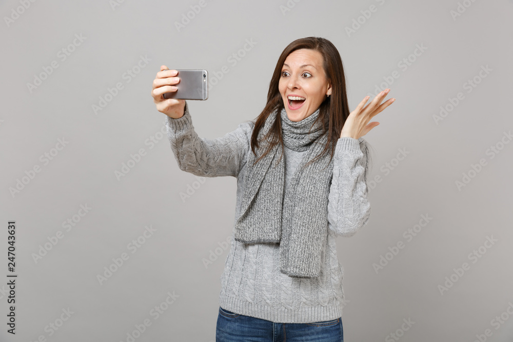 Obraz premium Excited young woman in sweater, scarf spreading hands, doing selfie shot on mobile phone, making video call isolated on grey background. Healthy fashion lifestyle people emotions cold season concept.