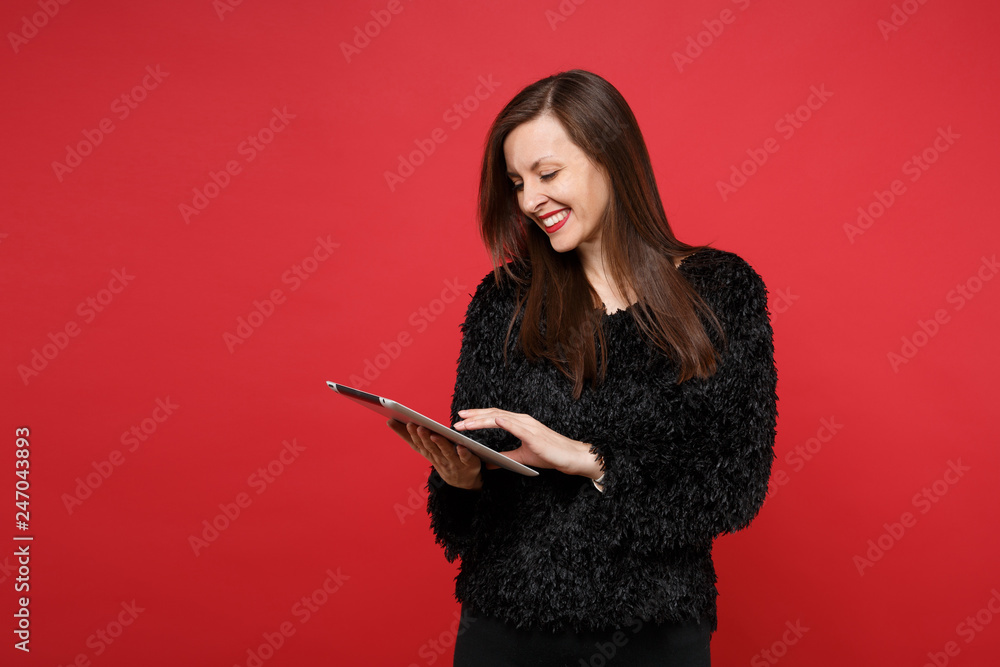 Portrait of stunning joyful young woman in black fur sweater using tablet pc computer isolated on bright red wall background in studio. People sincere emotions, lifestyle concept. Mock up copy space. Portrait of stunning joyful young woman in black fur sweater using tablet pc computer isolated on bright red wall background in studio. People sincere emotions, lifestyle concept. Mock up copy space.