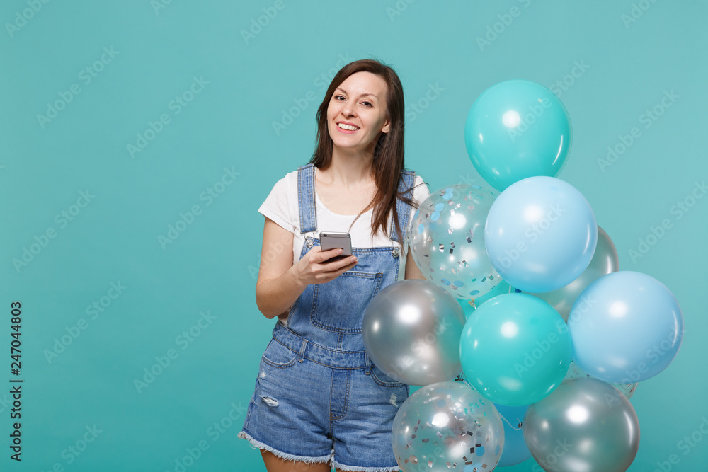 Fototapeta premium Beautiful smiling young woman in denim clothes holding mobile phone, celebrating with colorful air balloons isolated on blue turquoise wall background. Birthday holiday party, people emotions concept.