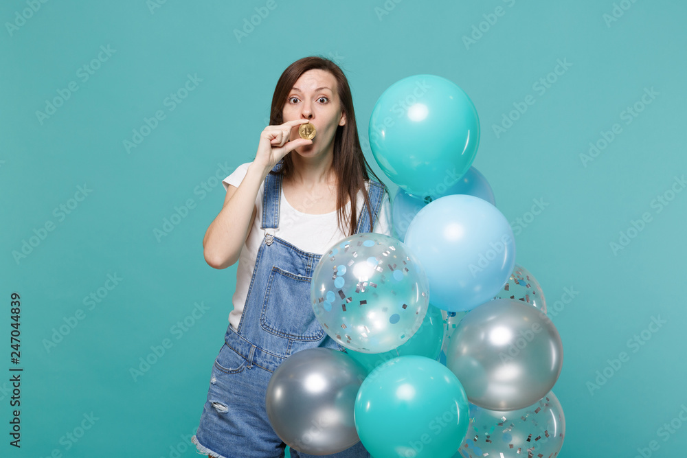 Fototapeta premium Amazed pretty young woman covering mouth with bitcoin, future currency celebrating holding colorful air balloons isolated on blue turquoise background. Birthday holiday party, people emotions concept.