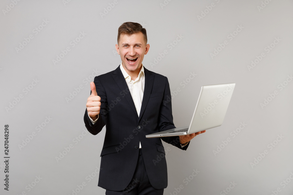 Joyful young business man in classic black suit shirt showing thumb up holding laptop pc computer isolated on grey background in studio. Achievement career wealth business concept. Mock up copy space. Joyful young business man in classic black suit shirt showing thumb up holding laptop pc computer isolated on grey background in studio. Achievement career wealth business concept. Mock up copy space.