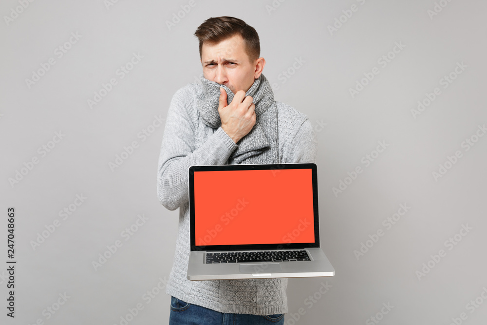 Young man in gray sweater covering mouth with scarf holding laptop pc computer with blank empty screen isolated on grey background. Healthy lifestyle, online treatment consulting, cold season concept. Young man in gray sweater covering mouth with scarf holding laptop pc computer with blank empty screen isolated on grey background. Healthy lifestyle, online treatment consulting, cold season concept.