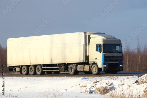 European white semi-trailer truck with logo copy space for writing on winter road on forest trees and blue sky background - side front view