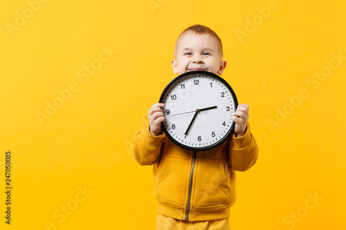 Little kid boy 3-4 years old wearing yellow clothes hold clock isolated on orange wall background, children studio portrait. People sincere emotions, childhood lifestyle concept. Mock up copy space.