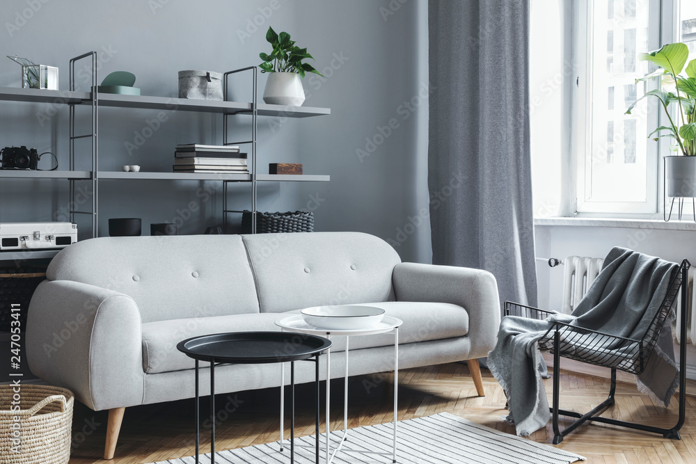 Modern and bright nordic living room with design sofa with pillow, coffee table, plants, stylish accessories and bookstand on the grey wall. Brown wooden parquet. Concept of minimalistic interior. Modern and bright nordic living room with design sofa with pillow, coffee table, plants, stylish accessories and bookstand on the grey wall. Brown wooden parquet. Concept of minimalistic interior.