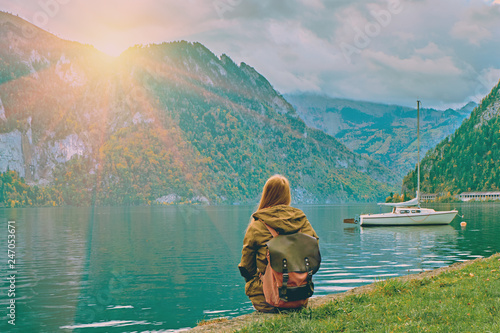Young tourist girl with backpack looking at the beautiful scenic sunrise at Austrian alps lake. Hipster travel vacation in alps mountains concept. Nice yacht boats on calm peaceful alps lake. Toned
