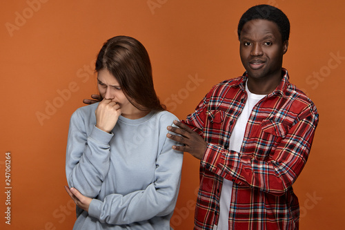 Human emotions, feelings and relationships concept. Caring supportive dark skinned young male dressed in plaid shirt embracing his emotional upset wife, saying words of support and encouragement