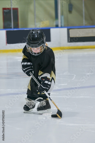 A little girl - hockey player on ice wearing in a full hockey equipment: helmet, black jersey, gloves with stick and puck.  