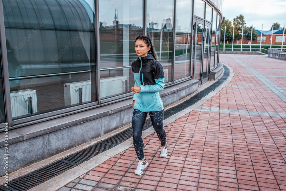 Girl athlete, summer city. Runs on a jog in a jump, playing sports on the street. In leggings and jacket. Free space for text. Sports in open air, evening run of the woman.