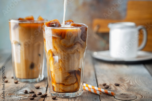 Ice coffee in a tall glass with cream poured over, coffee ice cubes and beans on a old rustic wooden table. Cold summer drink with tubes on a blue rusty background with copy space