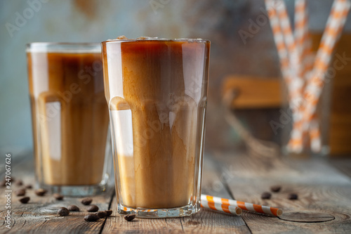 Ice coffee in a tall glass with cream poured over, coffee ice cubes and beans on a old rustic wooden table. Cold summer drink with tubes on a blue rusty background with copy space