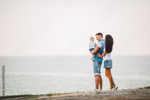 Family holiday in nature. Three, Mom, Dad, daughter one year standing with backs on cliff overlooking sea. man holding hild in arms,woman hugging husband.Fashionable people dressed in stylish clothes