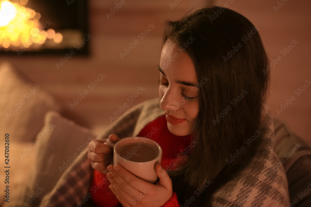 Young woman drinking coffee near decorative fireplace at home. Winter season Young woman drinking coffee near decorative fireplace at home. Winter season