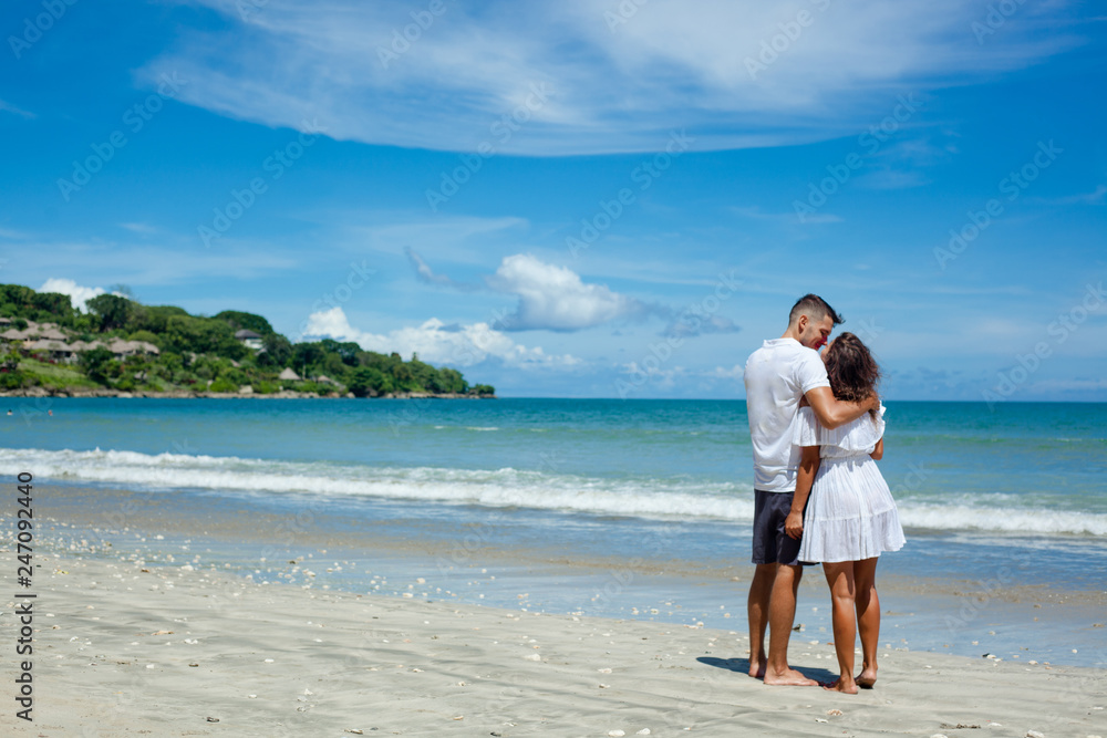 Back view of young romantic couple in white clothes standing on tropical beach. Man and woman huging on left side of photo. Blue sky and see on background Back view of young romantic couple in white clothes standing on tropical beach. Man and woman huging on left side of photo. Blue sky and see on background