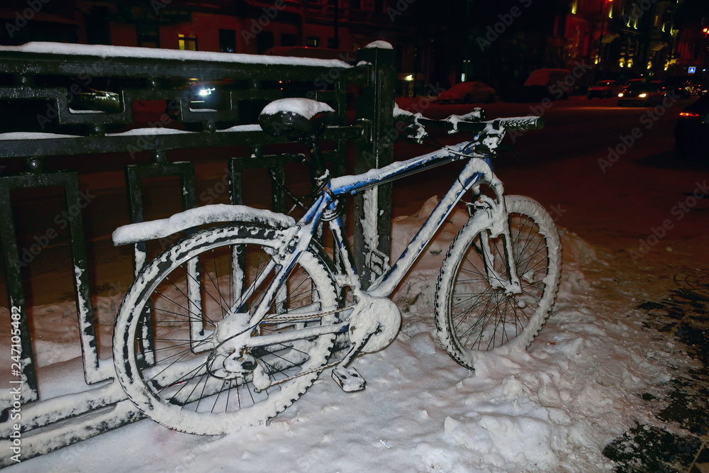 A snow-covered bike parked on a fence on a winter street.